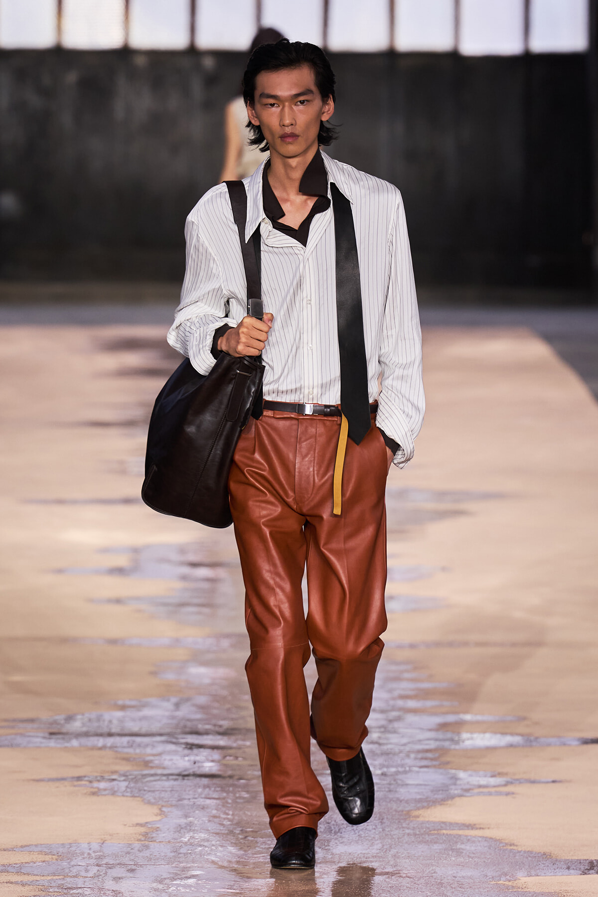 Male fashion model walking the runway in a white pinstriped shirt, brown leather pants, suspenders, and a black bag over the shoulder