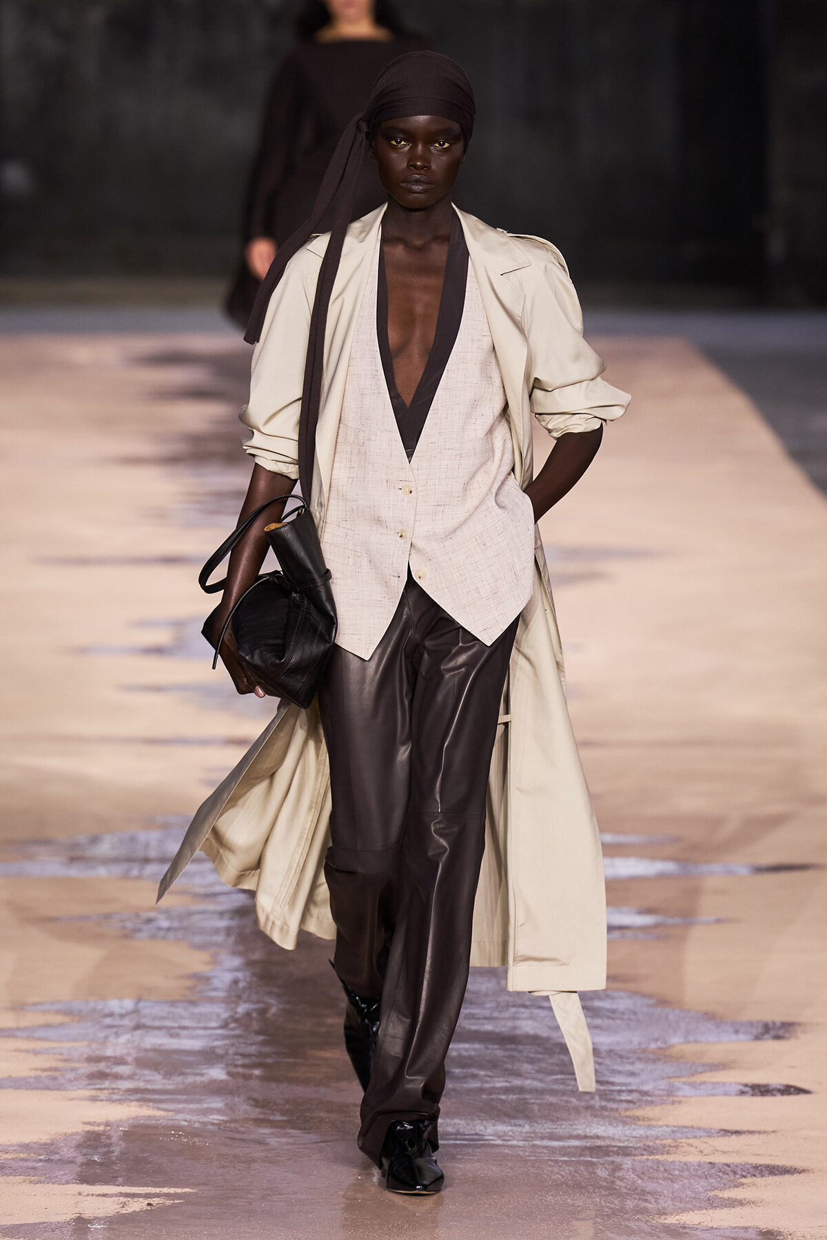 Dark-skinned model walking a runway in a beige trench coat over a light deep-V vest, paired with brown leather pants and a black headscarf, carrying a black bag.