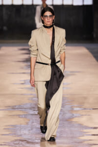 Female model walks a beige suit with a black draped scarf and belt on a runway, wearing sunglasses.