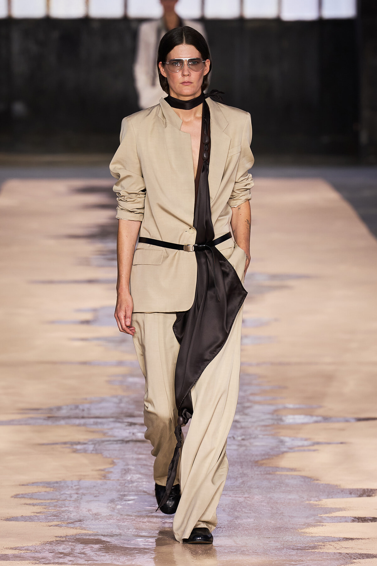 Female model walks a beige suit with a black draped scarf and belt on a runway, wearing sunglasses.