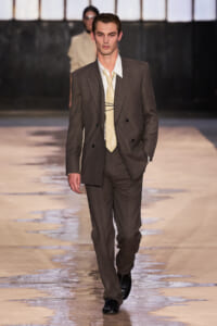Male model walking a runway in a brown double-breasted suit, white shirt, and pale yellow tie.