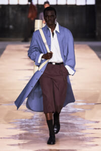 Male model on a runway wearing a light blue long coat over a white striped shirt and burgundy cropped trousers, with a beige shoulder strap and black loafers.