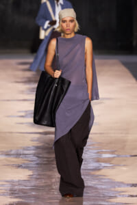Model walking the runway in a lavender, asymmetrical draped top with wide-leg black pants, a beige headscarf, and a large black tote.」