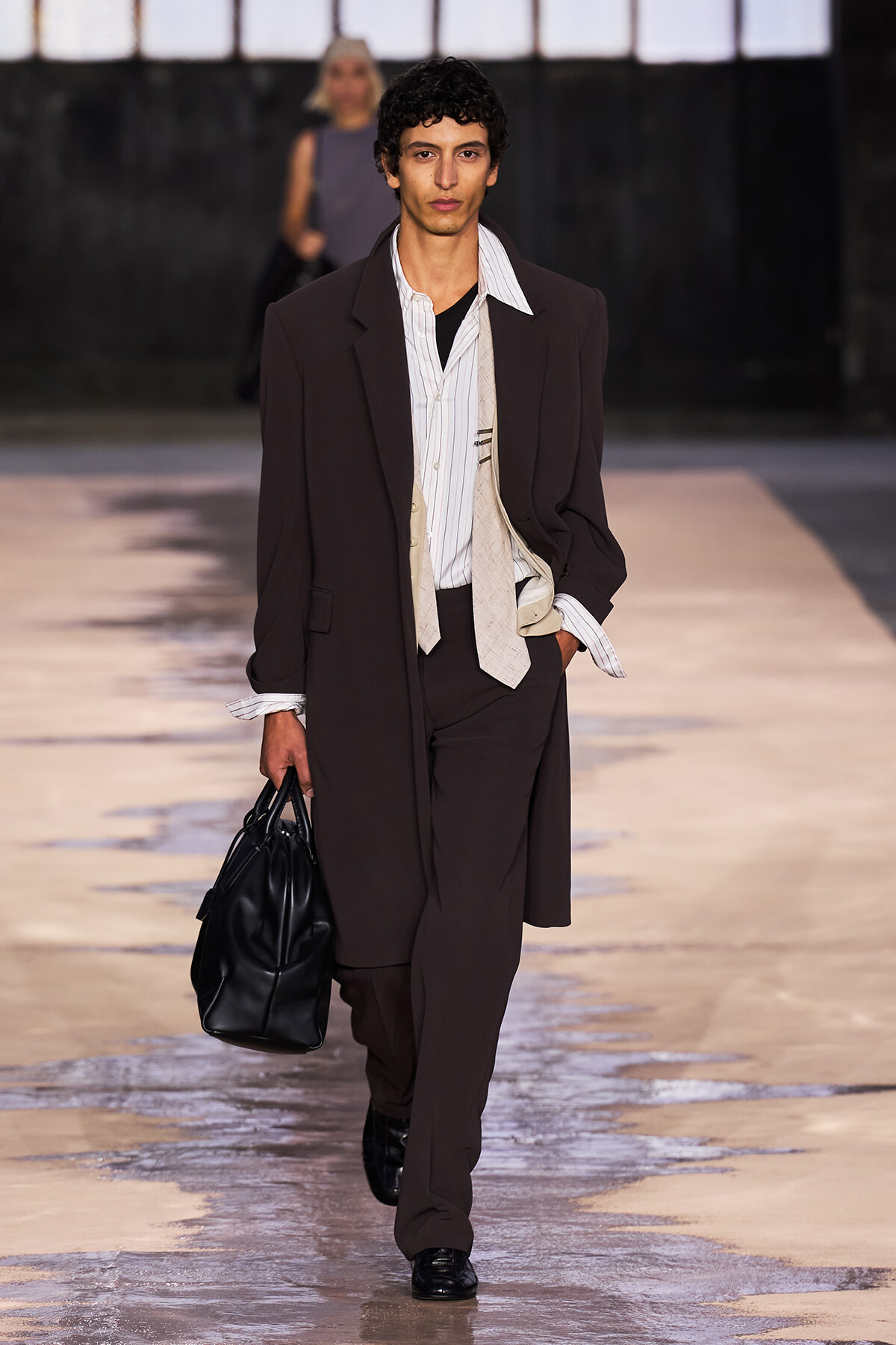 Male model walking a fashion runway in a dark brown suit, white striped shirt, loosely knotted beige tie, and a black leather bag in hand