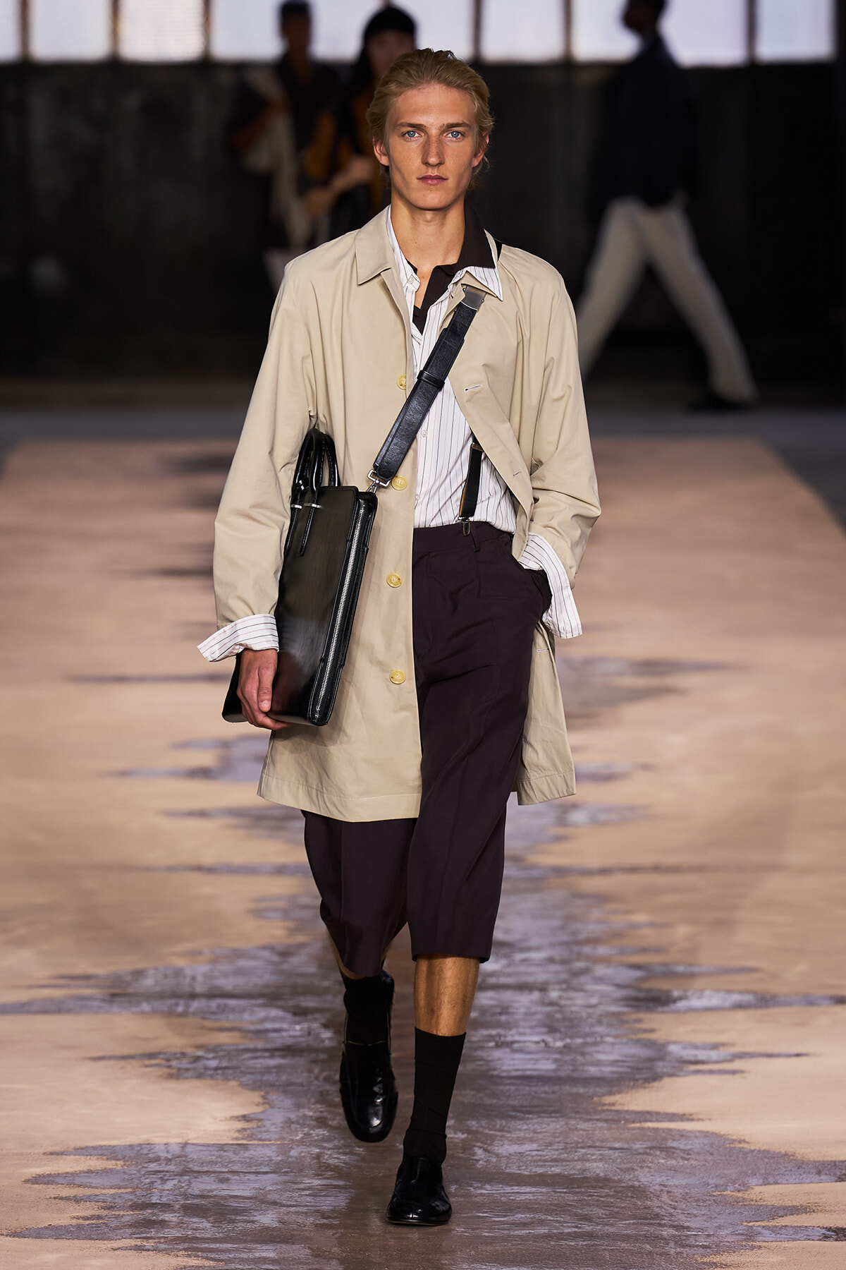 Male model walking a runway in a beige trench coat over a striped shirt, holding a black leather portfolio bag with a crossbody strap.