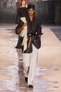 Model walks the runway in an oversized black blazer with a beige scarf draped over the shoulder, paired with light gray trousers and black shoes, carrying a cream bag.