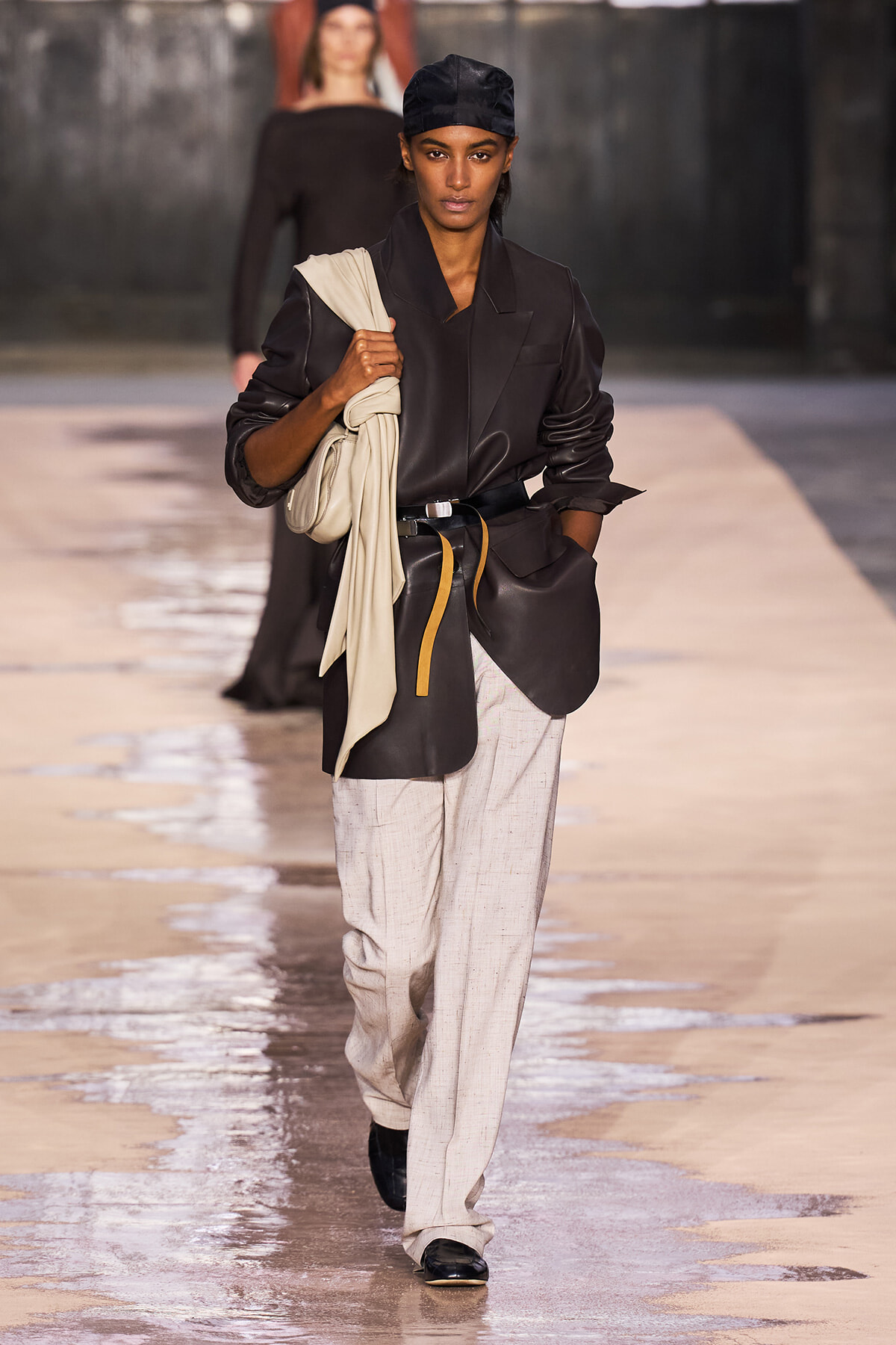 Model walks the runway in an oversized black blazer with a beige scarf draped over the shoulder, paired with light gray trousers and black shoes, carrying a cream bag.