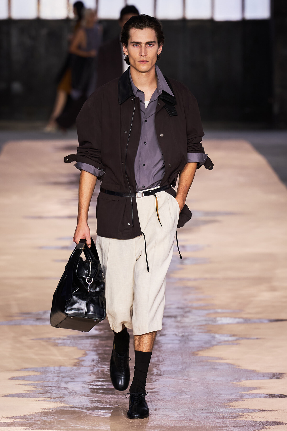 Male model on a runway wearing a brown jacket over a gray shirt, beige knee-length shorts, black shoes, and carrying a black leather bag.
