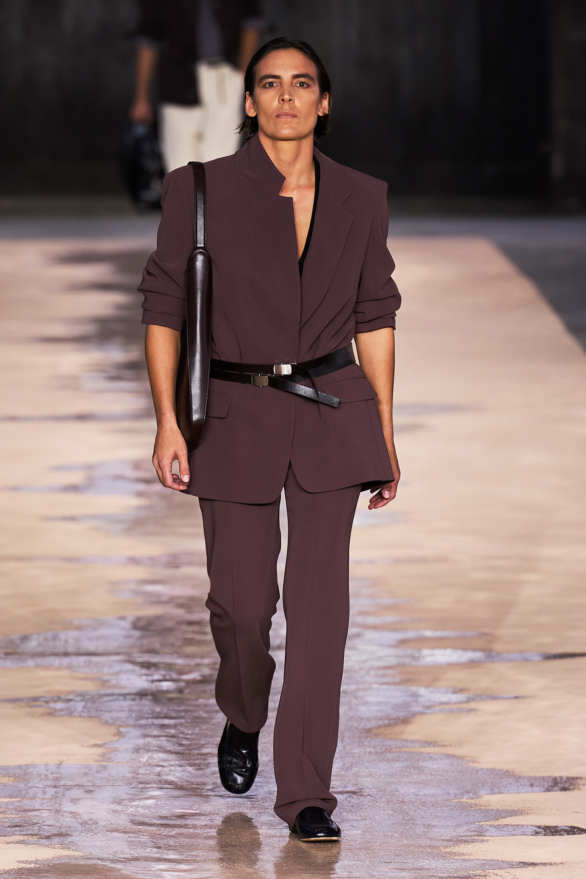 Model walks the runway in a burgundy tailored suit with a black belt and shoulder bag on a wet, reflective catwalk.