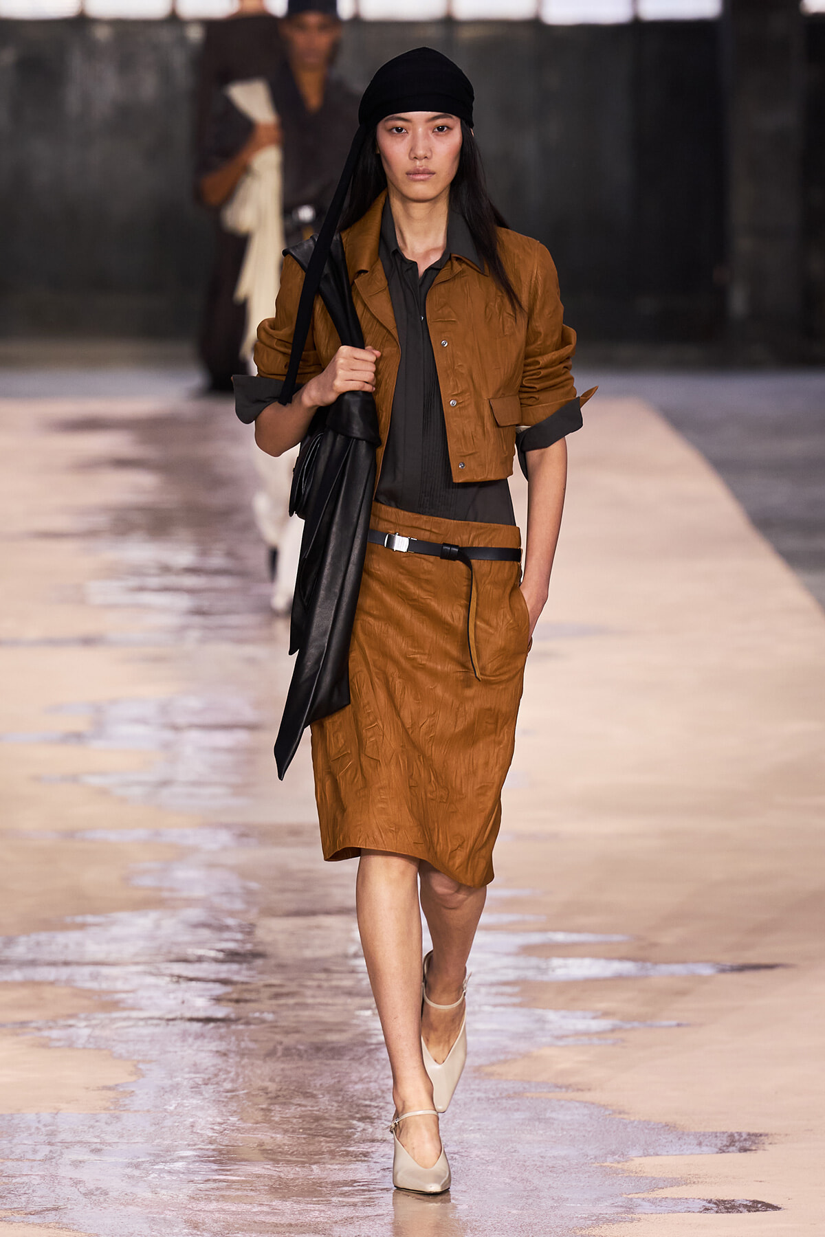 Model walks a runway in a brown jacket and skirt set, black top, black headscarf, and beige heels, carrying a black bag.