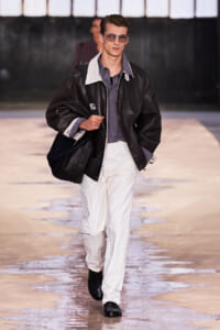 Male model walking runway in oversized black leather jacket, gray shirt, white pants, and sunglasses carrying a black bag.