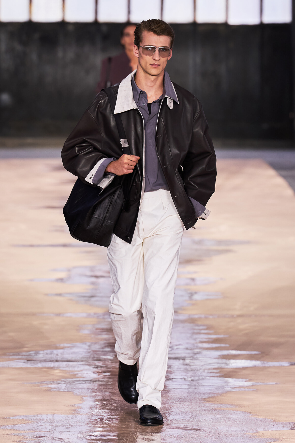 Male model walking runway in oversized black leather jacket, gray shirt, white pants, and sunglasses carrying a black bag.