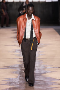 Male model walks the runway in an orange brown leather jacket over a white shirt and dark brown trousers.