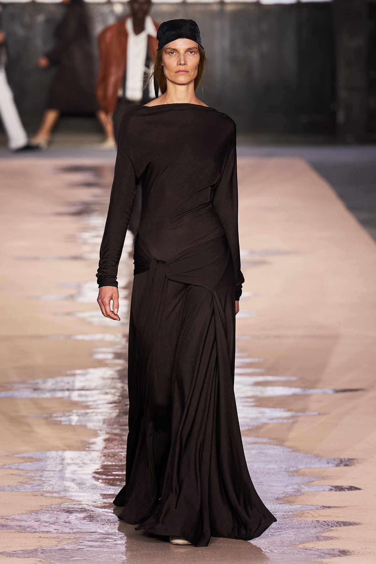Female model walks down a fashion runway in a long black draped gown and a black headscarf, spotlighted against a neutral backdrop.