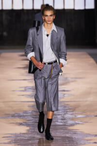 Male model walking a fashion runway in a light gray suit with a striped shirt, rolled cuffs, and black shoes; carrying a black bag over the shoulder.