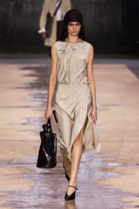 Model walking the runway in a beige draped sleeveless dress, black headwrap, and carrying a shiny black handbag.