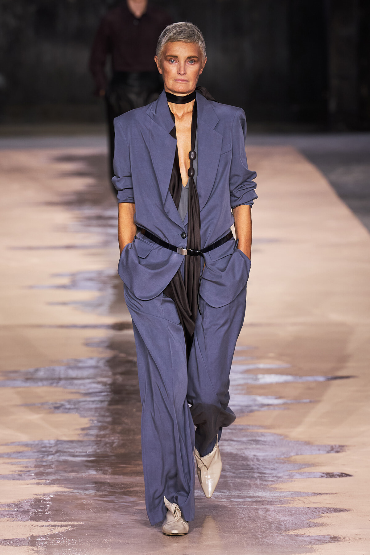 Model walks the runway in a loose blue suit with a plunging black top and cream boots, hands in pockets.