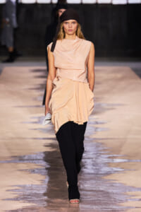 Model walking down a runway in a peach draped satin top, black pants, and a dark headscarf; holding a small white clutch.