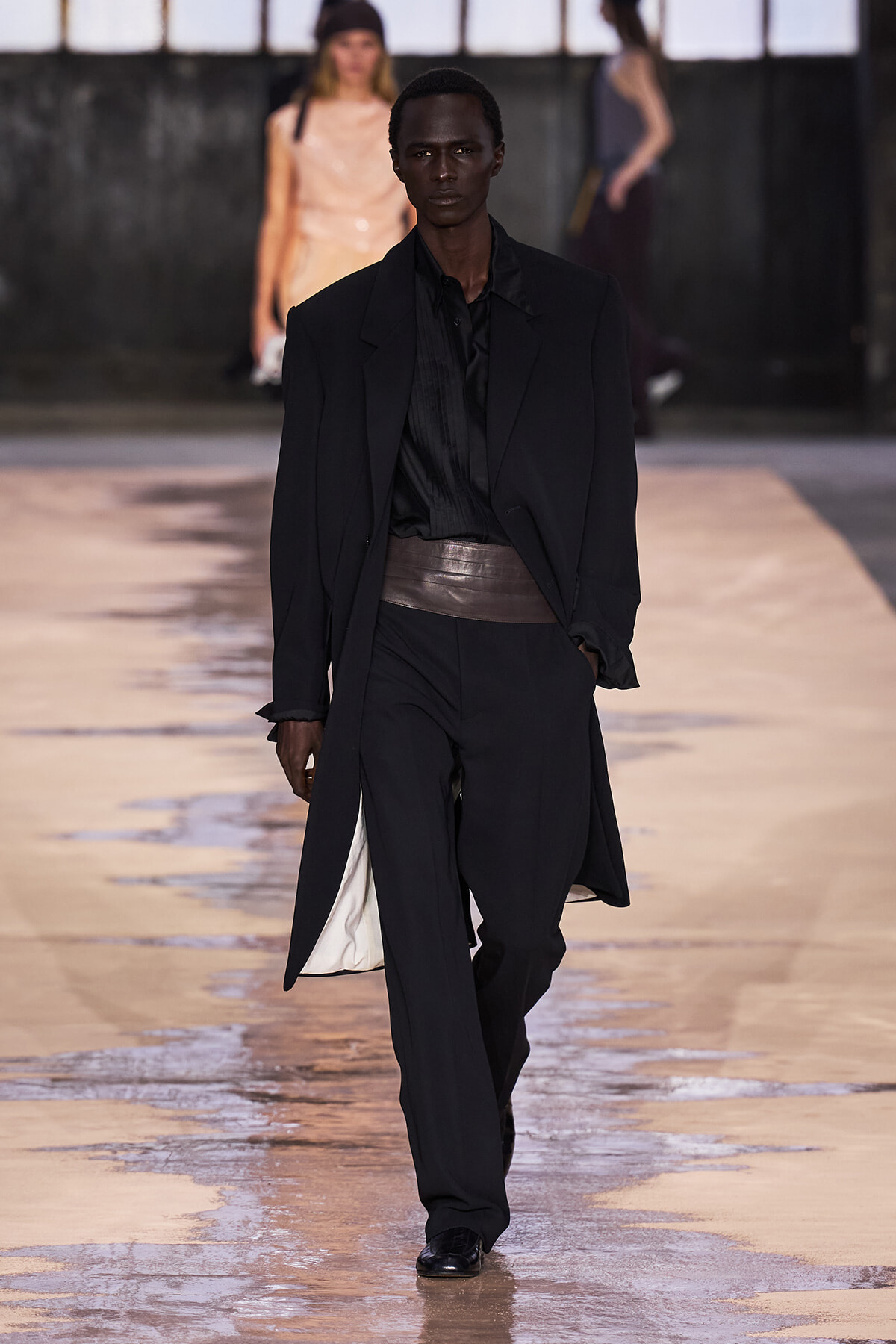 Male model walking a fashion runway in a black suit with a long coat and wide brown waist belt.