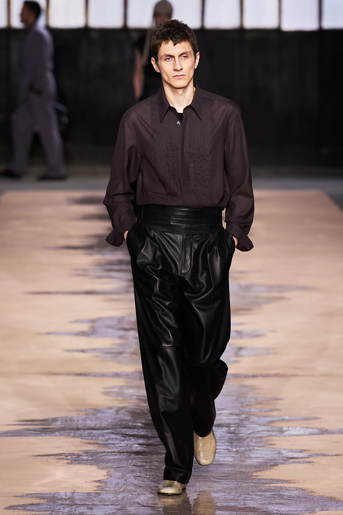 Male model walking a runway in a dark brown button-down shirt and high-waisted black leather pants.