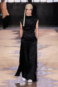 Model walks the runway in a long black satin dress with side pockets, wearing a beige headscarf and white sneakers.