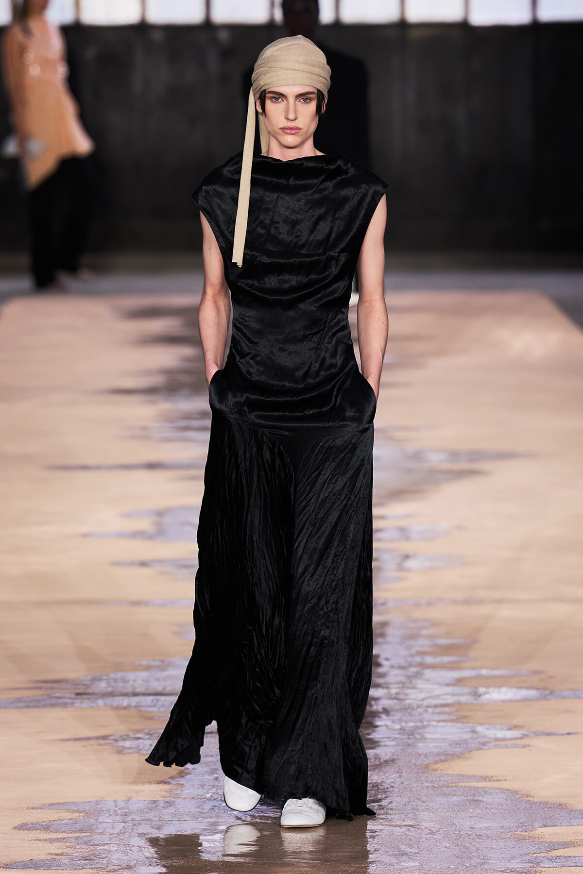 Model walks the runway in a long black satin dress with side pockets, wearing a beige headscarf and white sneakers.
