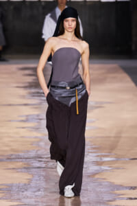 Model on a runway wearing a gray strapless top with oversized dark trousers, black head wrap, and belt.