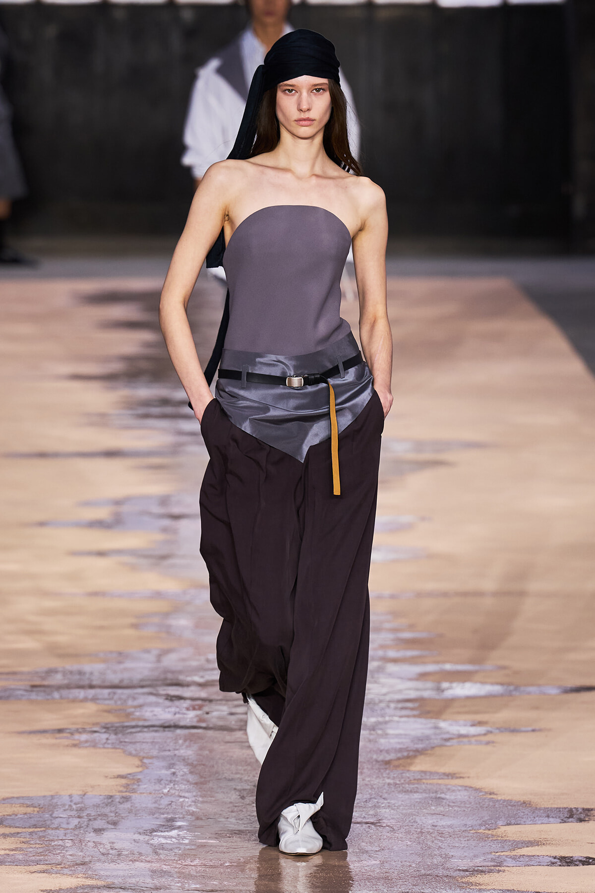 Model on a runway wearing a gray strapless top with oversized dark trousers, black head wrap, and belt.