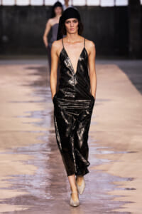 Model walking a fashion runway in a shiny black V-neck slip dress with a black headscarf and beige flats.