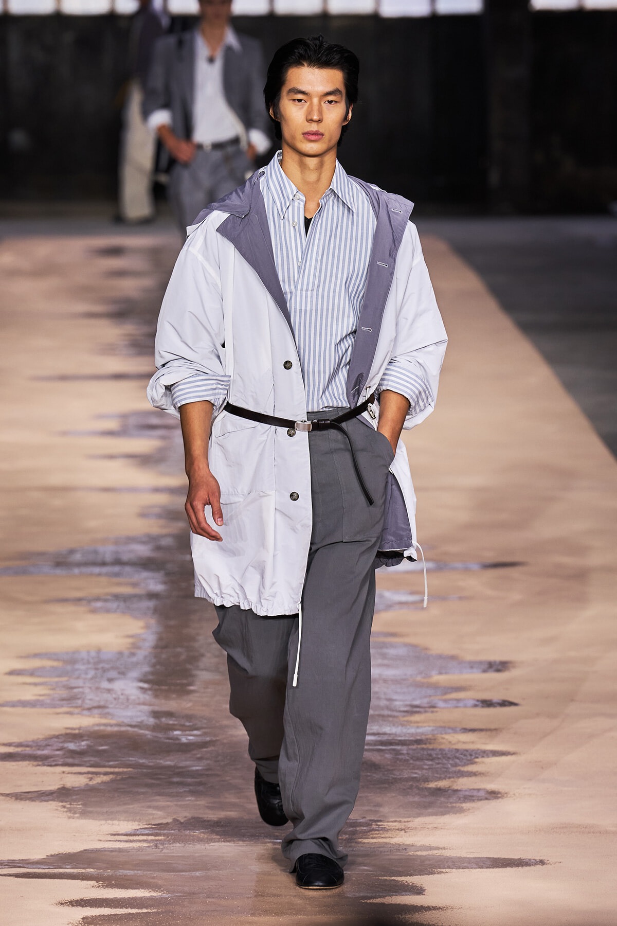 Male model walks a fashion runway in a light gray oversized jacket over a blue striped shirt and gray wide-leg trousers.