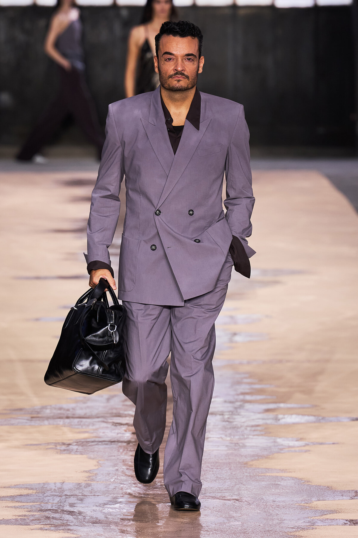Male model walking a runway in a lavender double-breasted suit, carrying a large black bag.