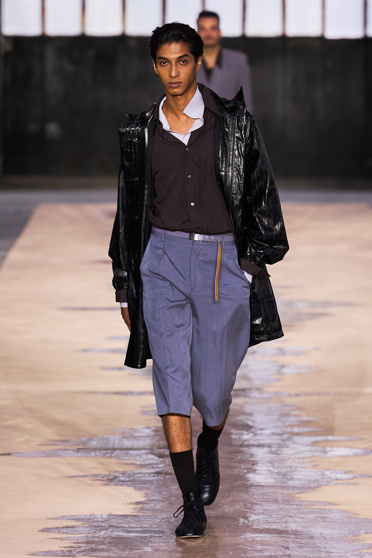 Male fashion model walking the runway in a glossy black coat over a brown shirt and light blue knee-length shorts with black boots and socks.