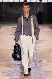 Male model walking a fashion show runway in a gray shirt, cream trousers with suspenders, carrying a black leather bag.