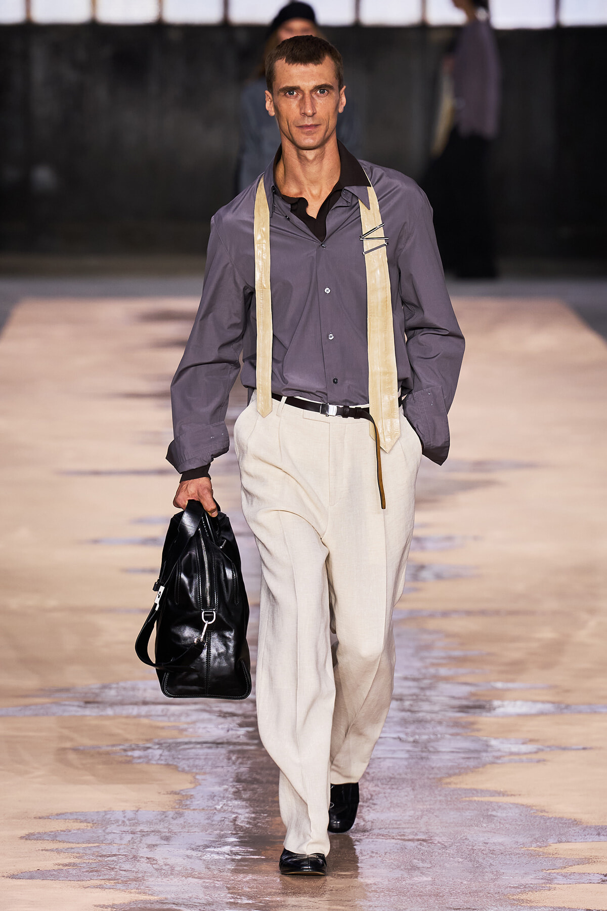Male model walking a fashion show runway in a gray shirt, cream trousers with suspenders, carrying a black leather bag.