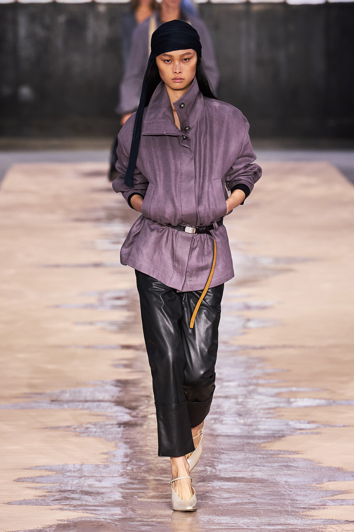 Model walks the runway in a lavender oversized jacket cinched with a thin belt, paired with black leather pants and beige heels, wearing a black headscarf.