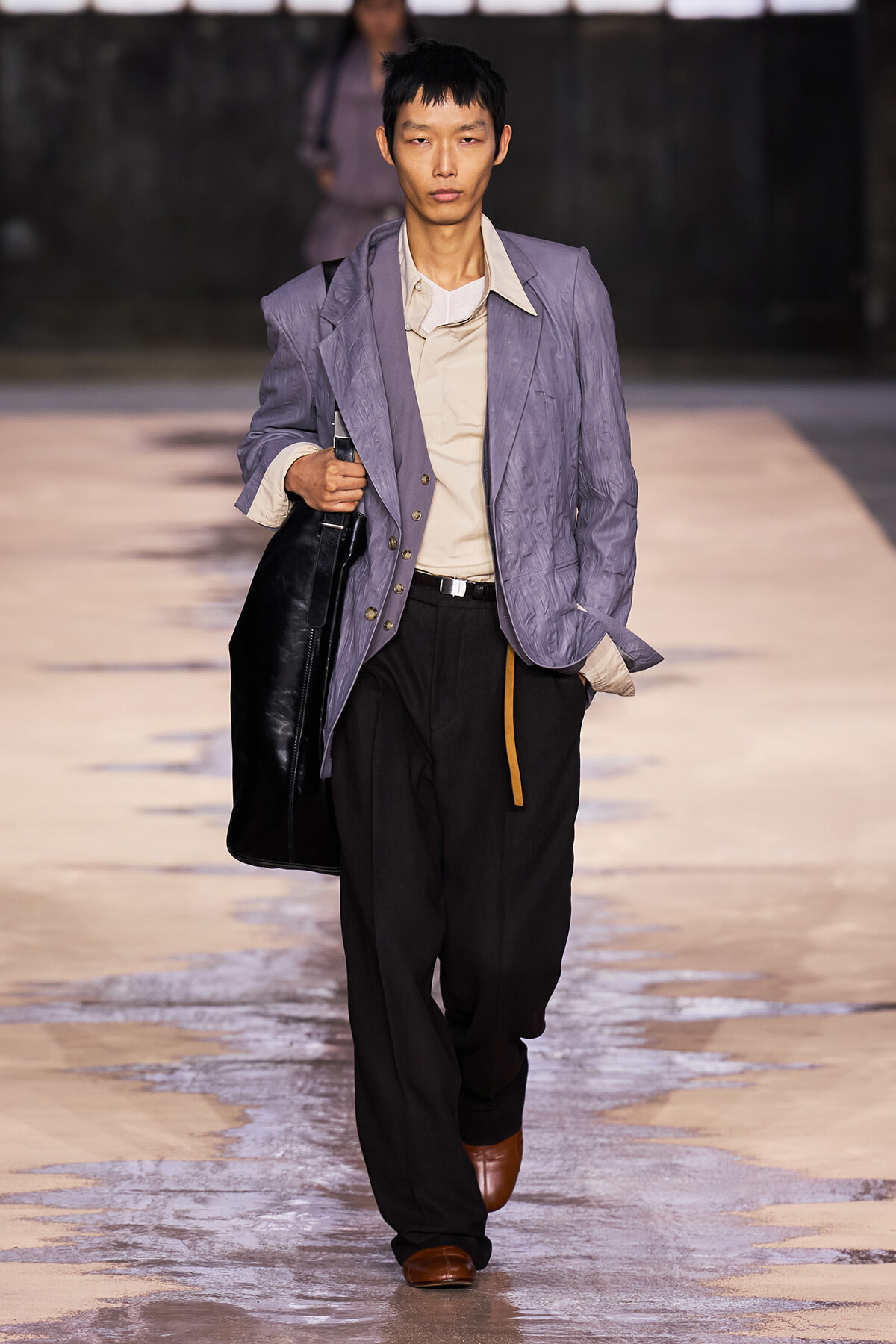 Male model walking a runway in a lavender quilted blazer over a beige shirt, black trousers, brown shoes, carrying a black bag on the shoulder/hand.