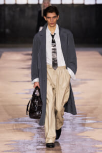 Male model on a runway wearing a long gray overcoat, striped shirt, metallic tie, and beige wide-leg pants, carrying a dark leather bag.