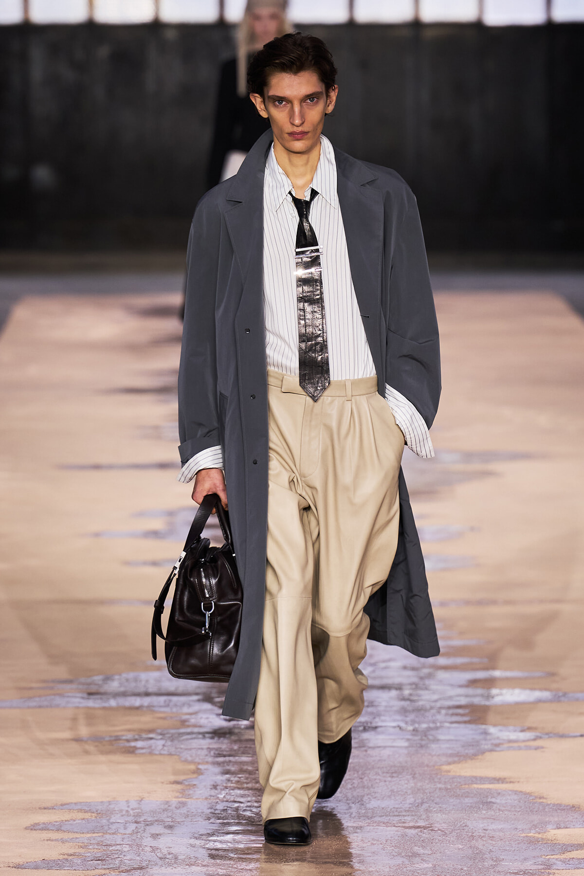Male model on a runway wearing a long gray overcoat, striped shirt, metallic tie, and beige wide-leg pants, carrying a dark leather bag.