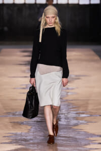 Model on runway wearing a black long-sleeve top with a light gray knee-length skirt featuring a beige panel, a tan headscarf, brown shoes, and a black handbag.