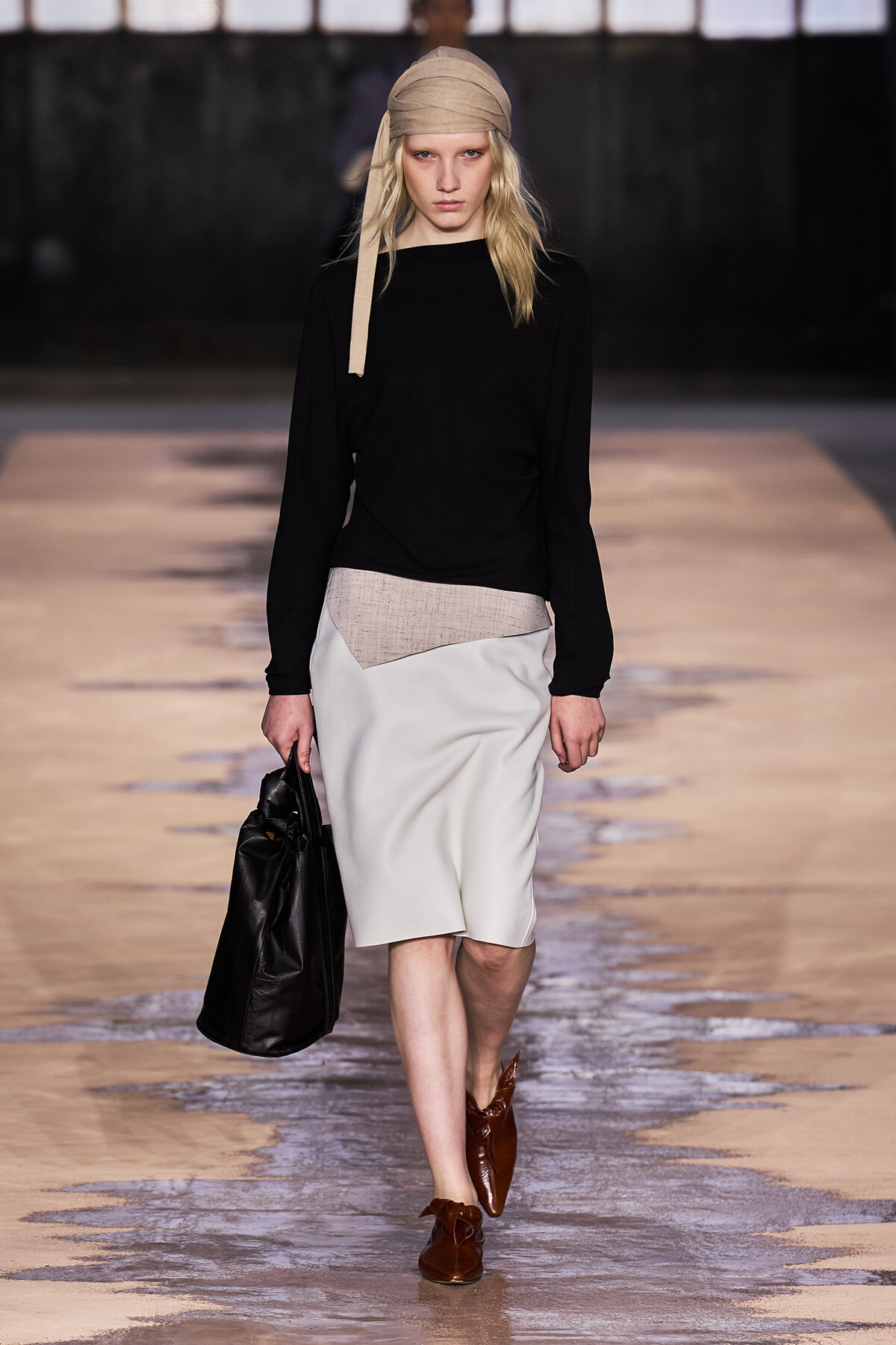 Model on runway wearing a black long-sleeve top with a light gray knee-length skirt featuring a beige panel, a tan headscarf, brown shoes, and a black handbag.