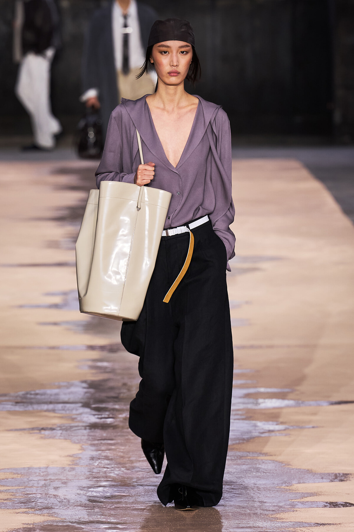 Model walks the runway in a loose purple blouse with a deep V, black wide-leg pants, and a large beige tote over the shoulder.