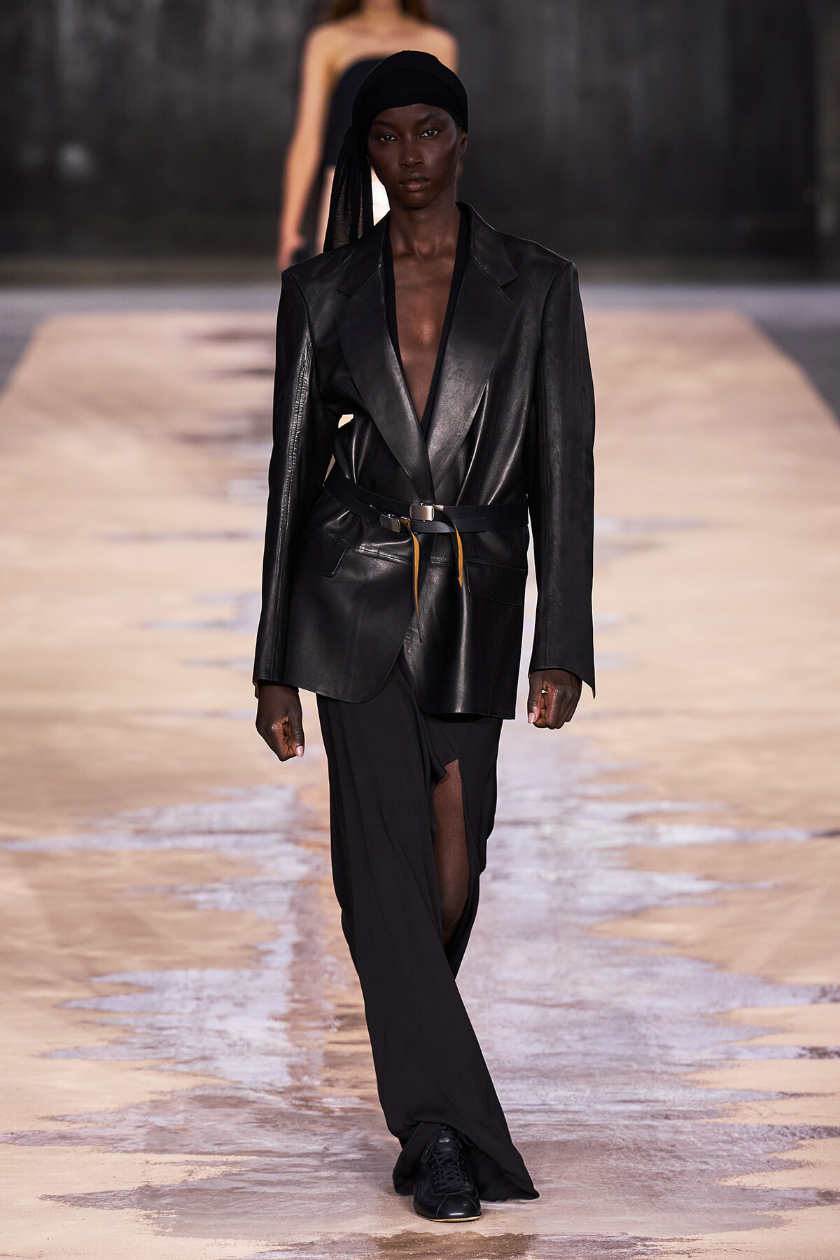 Model walks the runway in an all-black outfit: oversized black leather blazer belted at the waist and a flowing black skirt with a thigh-high slit, paired with a black head wrap.