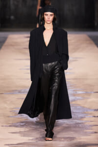 Female model walking down a runway in an all-black outfit: long blazer, deep V-neck top, leather pants, black headscarf, and open-toe sandals on a beige runway.