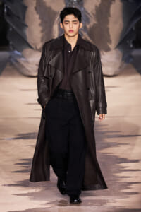 Male model walking a fashion runway in a long dark brown leather trench coat over black shirt and trousers; hands in pockets, serious expression, dramatic backdrop.