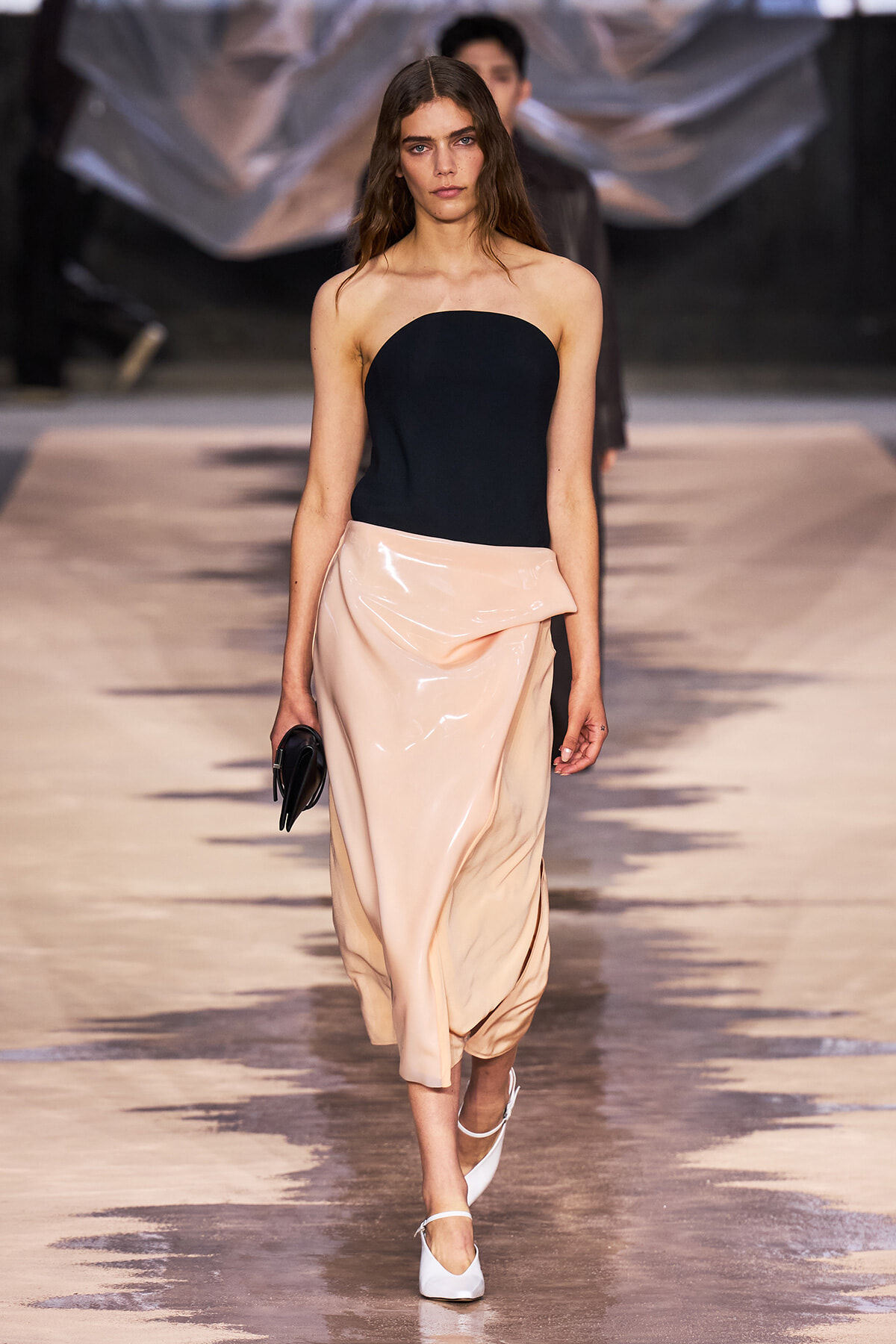 Model on a runway wearing a strapless black bodice and a pale pink satin draped skirt, carrying a black clutch.