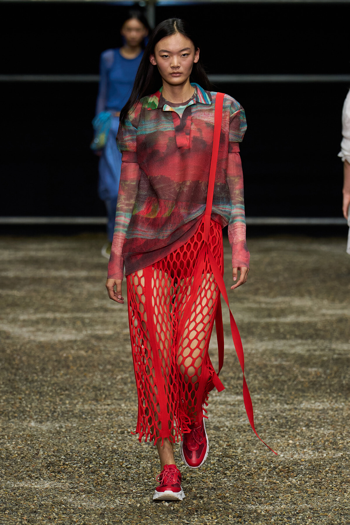 Model walking a runway in a sheer multicolor top and bold red fishnet skirt with matching red sneakers.
