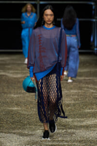Female model walking a runway in an oversized blue-grey top with red graphic, paired with a dark blue fishnet skirt and black sneakers, carrying a bright turquoise bag with a small black pouch.