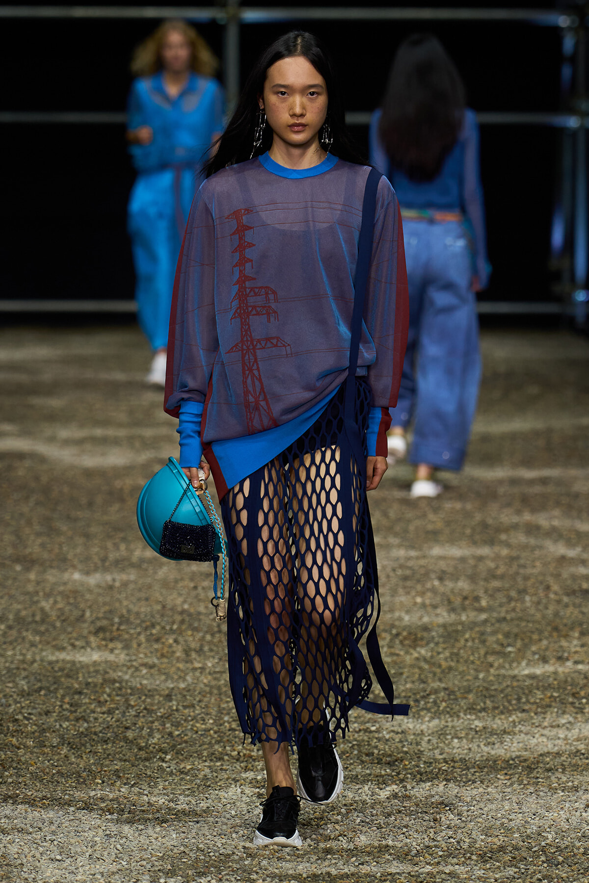 Female model walking a runway in an oversized blue-grey top with red graphic, paired with a dark blue fishnet skirt and black sneakers, carrying a bright turquoise bag with a small black pouch.