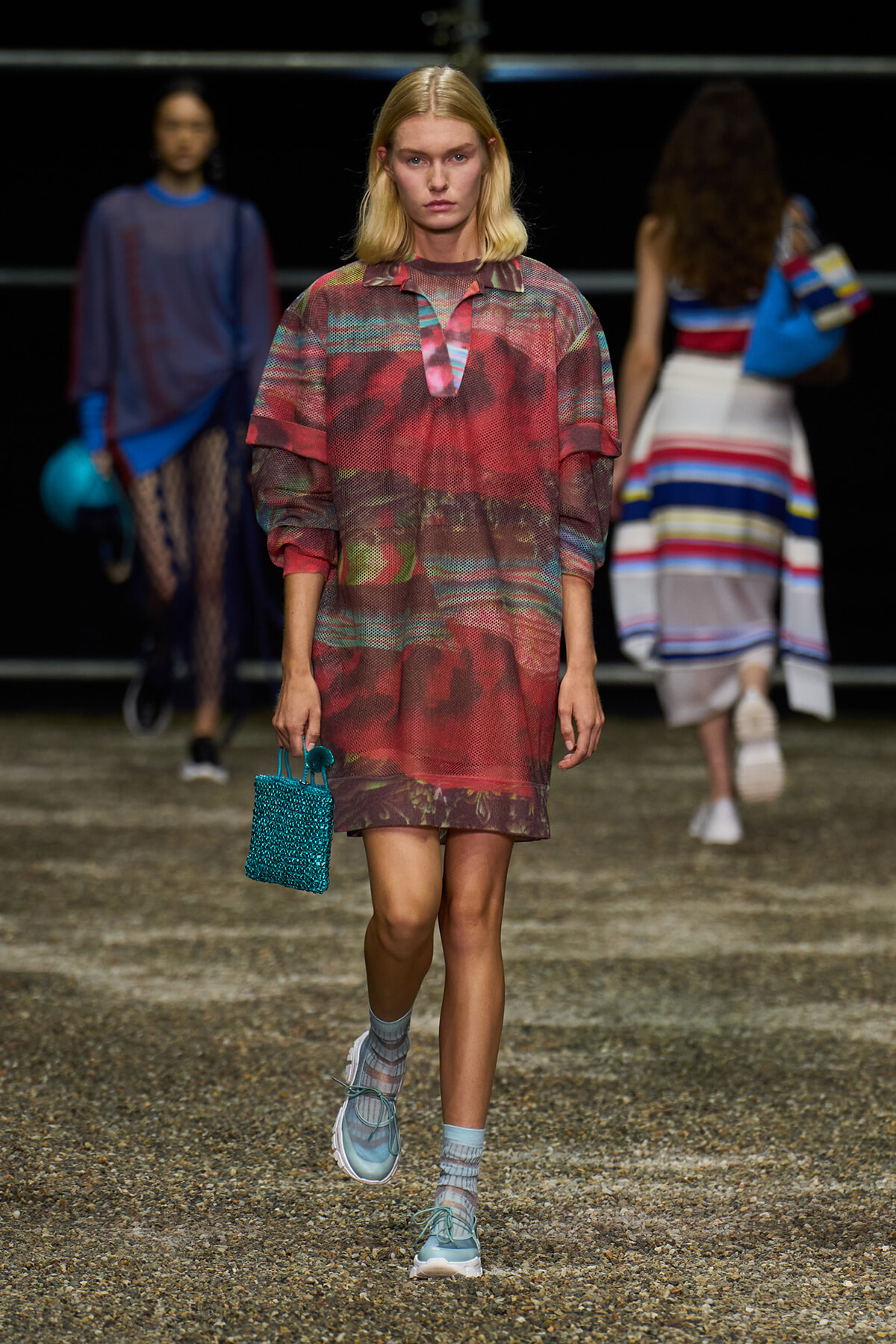 Model walking a fashion show runway in a red patterned shift dress, turquoise bag in hand, and light blue socks with sneakers.