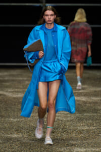Model walks a blue monochrome outfit down the runway, clutching a brown textured tote.